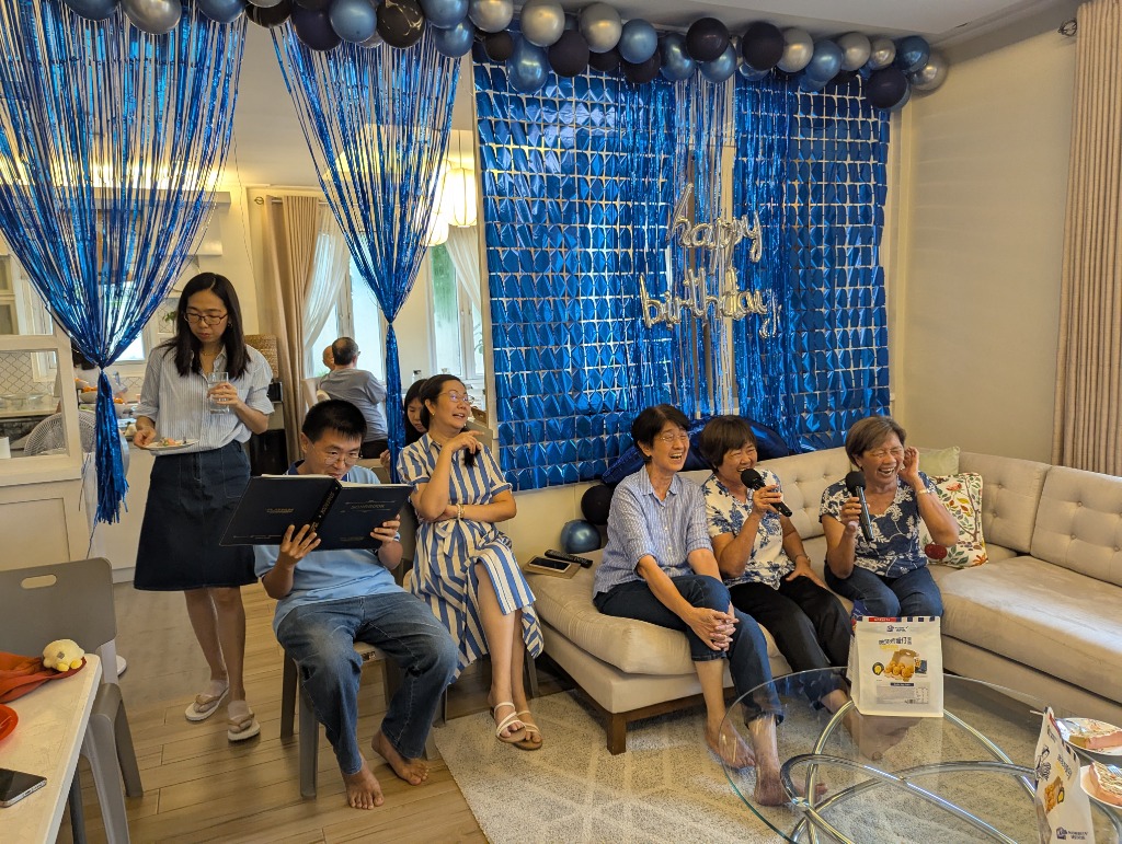 Wide view of the living room karaoke party with family singing, songbook, and festive blue backdrop