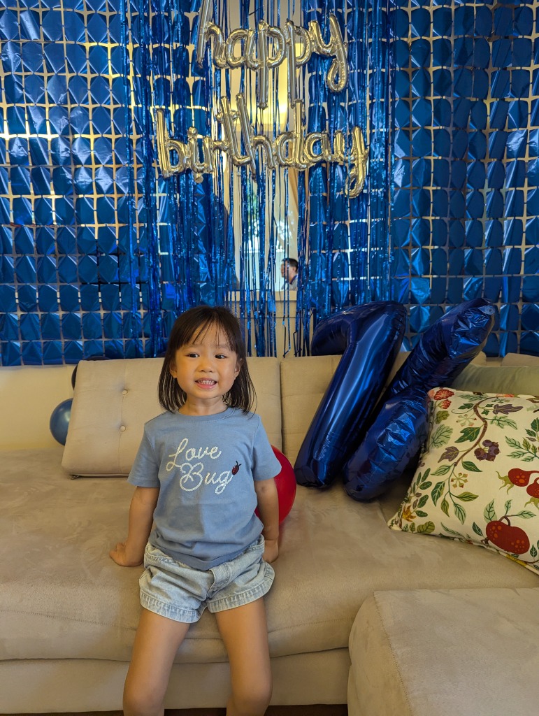 Little girl in blue Love Bug shirt smiling beside the 76 milestone balloons and blue metallic backdrop
